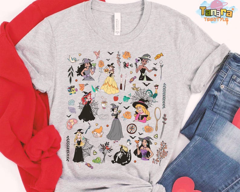 May include: A light gray t-shirt featuring a colorful Halloween-themed graphic. The design includes illustrations of witches, pumpkins, bats, and other spooky elements. The shirt is paired with blue jeans and a red sweatshirt.