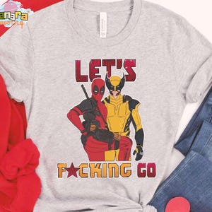 Puede incluir: Camiseta gris claro con ilustraciones de Deadpool y Wolverine. El texto "LET'S F*CKING GO" est&aacute; impreso en rojo y naranja. La camiseta se combina con vaqueros azules y una camisa roja de manga larga.