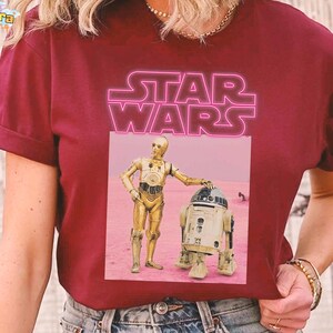 May include: Burgundy t-shirt featuring a pink neon "STAR WARS" logo. Below the logo is a graphic of C-3PO and R2-D2 standing on a pink surface. The t-shirt has short sleeves and a crew neck.