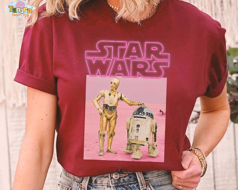 May include: Burgundy t-shirt featuring a pink neon "STAR WARS" logo. Below the logo is a graphic of C-3PO and R2-D2 standing on a pink surface. The t-shirt has short sleeves and a crew neck.