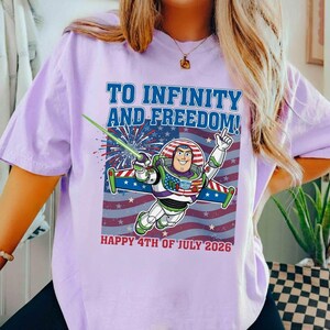 May include: A light purple t-shirt featuring a graphic of Buzz Lightyear with the text "TO INFINITY AND FREEDOM!" and "HAPPY 4TH OF JULY 2026" in blue and red. The design incorporates an American flag motif.