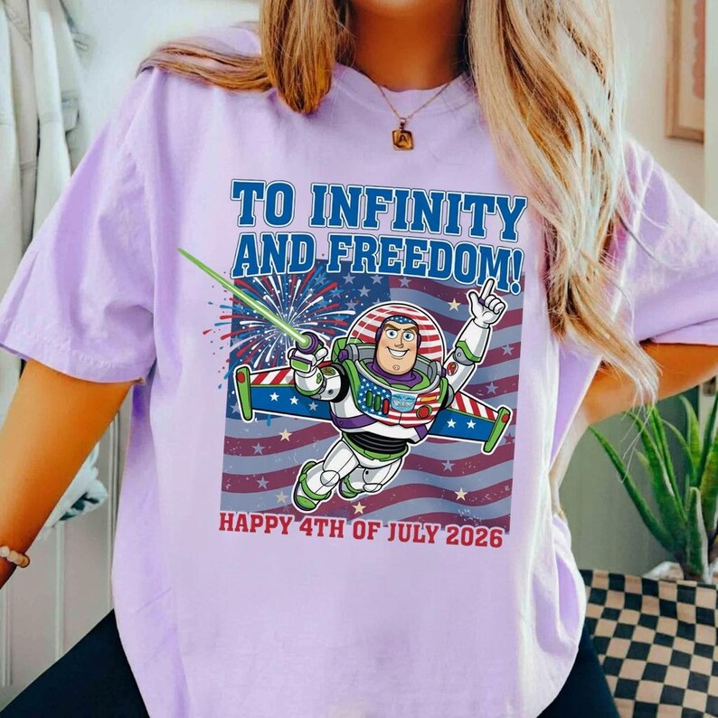May include: A light purple t-shirt featuring a graphic of Buzz Lightyear with the text "TO INFINITY AND FREEDOM!" and "HAPPY 4TH OF JULY 2026" in blue and red. The design incorporates an American flag motif.