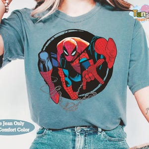 May include: A blue jean colored t-shirt featuring a graphic of Spider-Man in a dynamic pose. The superhero is depicted in his classic red and blue suit, set against a black circular background. The shirt also has the text "Blue Jean Only With Comfort Color".
