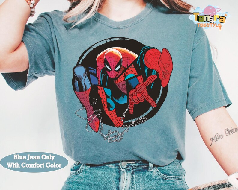 May include: A blue jean colored t-shirt featuring a graphic of Spider-Man in a dynamic pose. The superhero is depicted in his classic red and blue suit, set against a black circular background. The shirt also has the text "Blue Jean Only With Comfort Color".