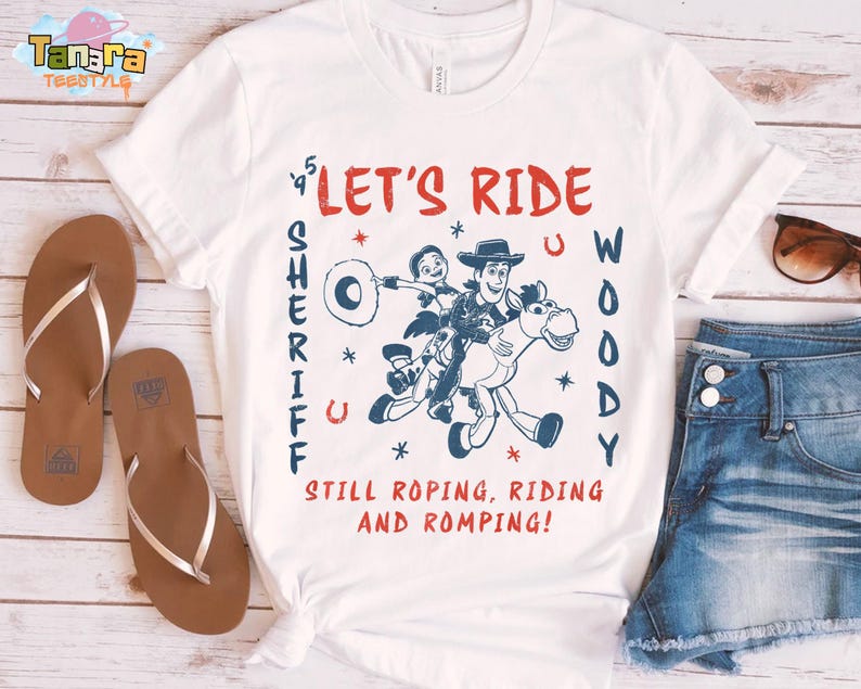 May include: White t-shirt with a graphic of Woody and Buzz Lightyear riding a horse, with the text "LET'S RIDE WOODY SHERIFF" in red and blue. The shirt also has the text "STILL ROPING, RIDING AND ROMPING!".