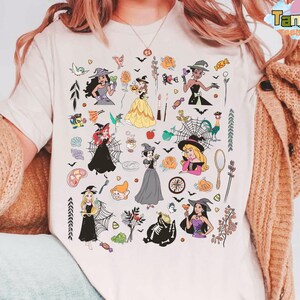 May include: Cream-colored t-shirt featuring a colorful graphic of various cartoon characters in Halloween costumes. The design includes bats, spiders, and other spooky elements. The shirt has a crew neck and short sleeves.