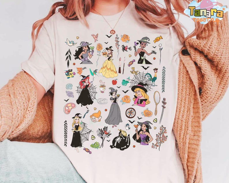 May include: Cream-colored t-shirt featuring a colorful graphic of various cartoon characters in Halloween costumes. The design includes bats, spiders, and other spooky elements. The shirt has a crew neck and short sleeves.