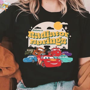 Pode incluir: T-shirt preto com a frase "Welcome to Radiator Springs" em amarelo e laranja, com desenhos de carros. O design inclui um sol e estrelas. A t-shirt tem gola redonda.