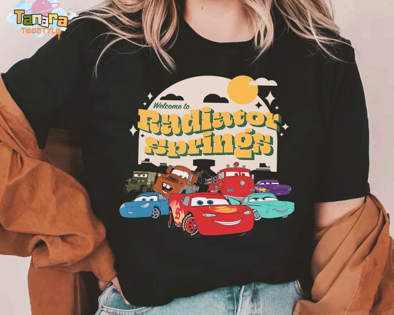 Pode incluir: T-shirt preto com a frase "Welcome to Radiator Springs" em amarelo e laranja, com desenhos de carros. O design inclui um sol e estrelas. A t-shirt tem gola redonda.