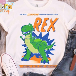 Puede incluir: Camiseta blanca con una ilustraci&oacute;n de dibujos animados de un dinosaurio. El dise&ntilde;o presenta un dinosaurio verde llamado Rex, con el texto "THE MOST TERRIFYING DINOSAUR WHO EVER LIVED!" y "AVAILABLE AT TOY STORES EVERYWHERE!" en naranja y azul.