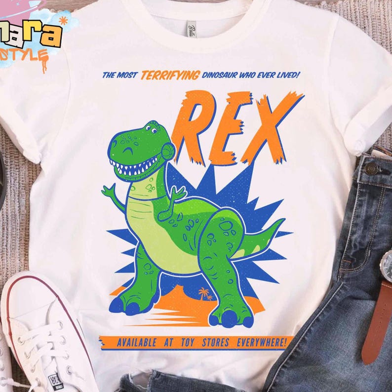 Puede incluir: Camiseta blanca con una ilustraci&oacute;n de dibujos animados de un dinosaurio. El dise&ntilde;o presenta un dinosaurio verde llamado Rex, con el texto "THE MOST TERRIFYING DINOSAUR WHO EVER LIVED!" y "AVAILABLE AT TOY STORES EVERYWHERE!" en naranja y azul.