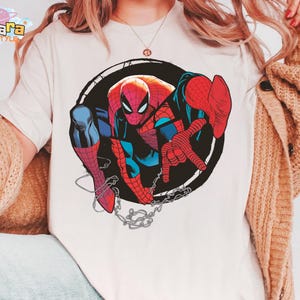 May include: White t-shirt featuring a colorful illustration of Spider-Man within a black circular frame. The superhero is depicted in his classic red and blue suit, with a web design and chain details. The shirt has the brand name "Tanara TEESTYLE" on the top left.