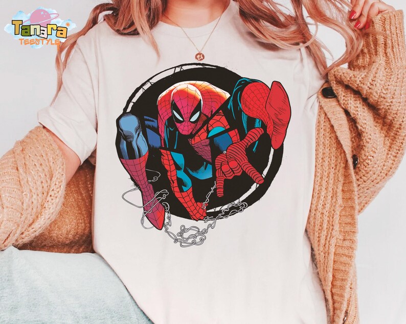 May include: White t-shirt featuring a colorful illustration of Spider-Man within a black circular frame. The superhero is depicted in his classic red and blue suit, with a web design and chain details. The shirt has the brand name "Tanara TEESTYLE" on the top left.