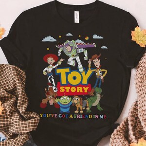 May include: Black t-shirt featuring a colorful graphic of characters from the animated film "Toy Story." The design includes Buzz Lightyear, Woody, Jessie, and other characters, with the words "Toy Story" and "You've Got a Friend in Me."