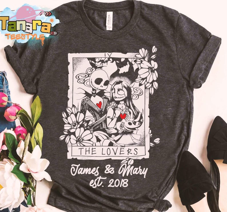 May include: Dark gray t-shirt featuring a black and white illustration of Jack Skellington and Sally from The Nightmare Before Christmas, with the text "THE LOVERS" and "James & Mary est. 2018". The design includes floral elements and red heart details.