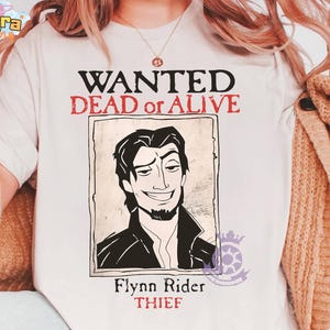 Puede incluir: Camiseta color crema con un diseño de cartel de "Se busca". El cartel tiene un retrato de un hombre con el texto "WANTED DEAD or ALIVE" en rojo y negro. Debajo del retrato, se lee "Flynn Rider THIEF".
