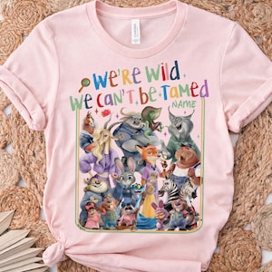 May include: A light pink t-shirt featuring a colorful graphic of various cartoon animals. The text "WE'RE WILD WE CAN'T BE TAMED" is displayed above the characters. The shirt is made of a soft material.