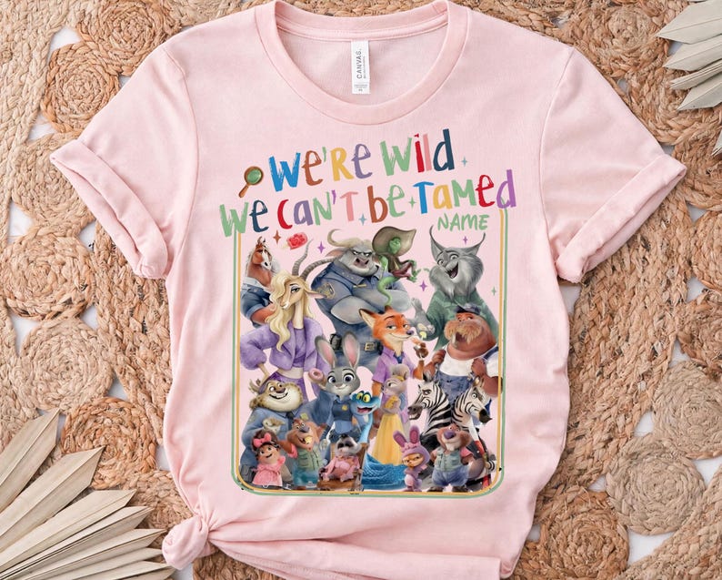 May include: A light pink t-shirt featuring a colorful graphic of various cartoon animals. The text "WE'RE WILD WE CAN'T BE TAMED" is displayed above the characters. The shirt is made of a soft material.