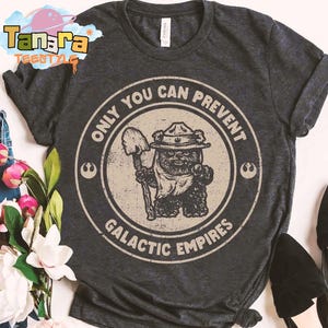 May include: Dark gray t-shirt with a circular graphic featuring an Ewok character holding a shovel, wearing a hat, and the text "ONLY YOU CAN PREVENT GALACTIC EMPIRES." The shirt has a crew neck and short sleeves. The shirt is displayed with a pair of black flats and a bouquet of flowers.