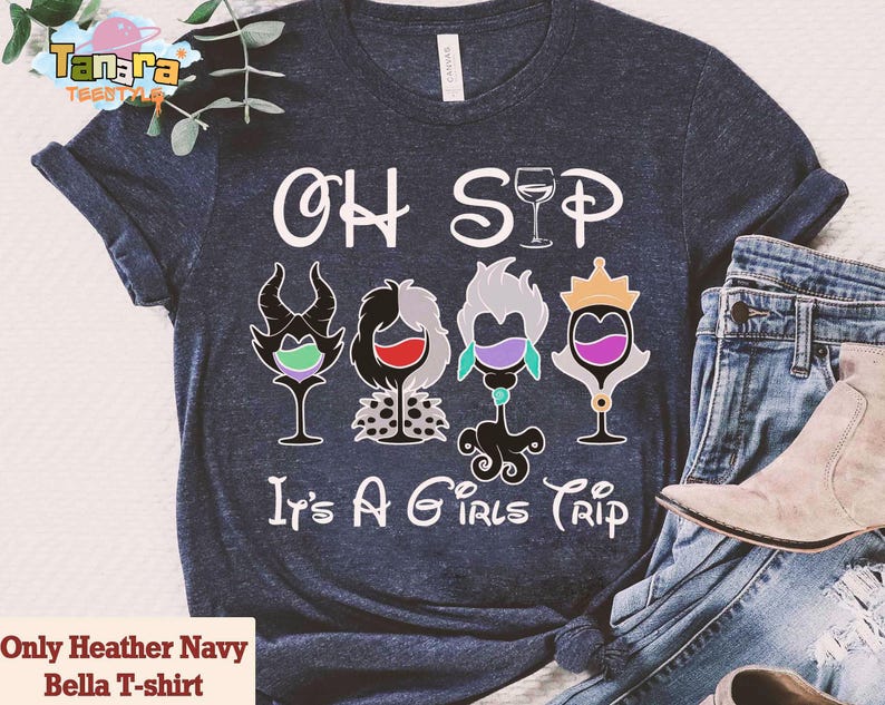 May include: Heather navy Bella T-shirt with the text "OH SIP It's A GIRLS TRIP". The shirt features four wine glass designs with cartoon character elements. The shirt is paired with blue jeans and a beige boot.