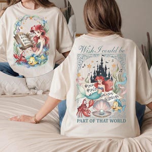 May include: Cream-colored t-shirt featuring a colorful illustration of a mermaid, crab, and fish, with the text "Wish I could be part of that world." The front of the shirt has a similar design.