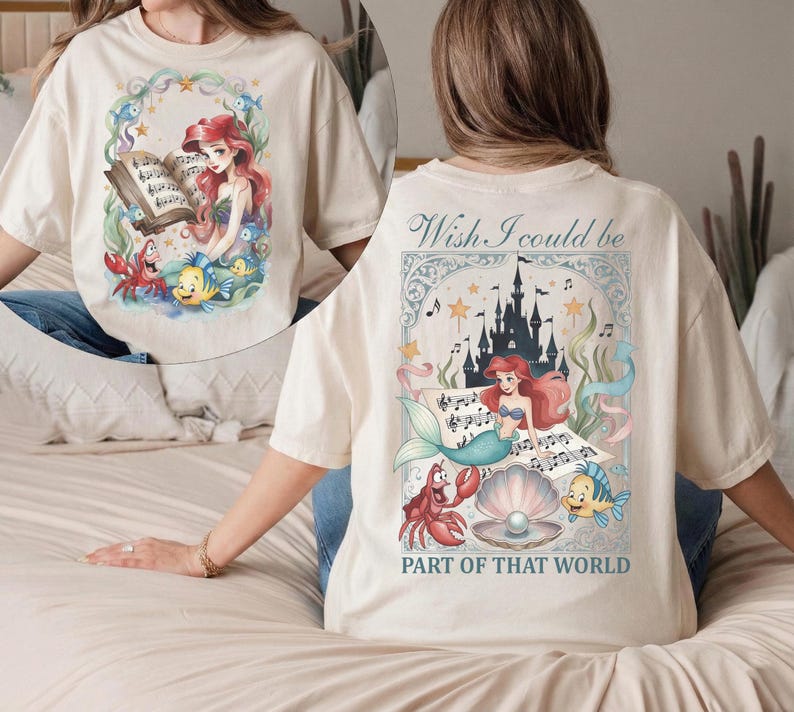 May include: Cream-colored t-shirt featuring a colorful illustration of a mermaid, crab, and fish, with the text "Wish I could be part of that world." The front of the shirt has a similar design.