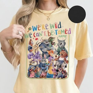 May include: A light yellow t-shirt featuring a colorful graphic of various cartoon characters. The text "We're Wild We Can't Be Tamed" is displayed above the characters. The shirt is made of a soft material.