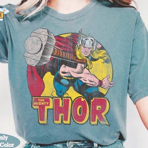 May include: A teal-colored t-shirt featuring a vintage comic book illustration of Thor, wielding his hammer. The graphic includes a yellow background, a red circle, and the text "THE MIGHTY THOR" in bold, orange letters. The shirt has short sleeves.