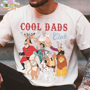 May include: Cream-colored t-shirt featuring the text "COOL DADS Club" in red and blue. The shirt displays cartoon characters including Mr. Incredible, Goofy, and Simba. A fun, graphic tee.