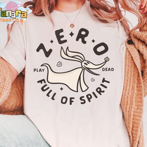 Puede incluir: Camiseta color crema con un gr&aacute;fico circular. El gr&aacute;fico presenta el texto "ZERO" sobre un personaje de dibujos animados parecido a un perro y las palabras "PLAY DEAD FULL OF SPIRIT" debajo. La camiseta es parte de la colecci&oacute;n Tanara TEENSTYLE.