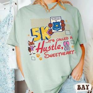 May include: A light green t-shirt with a graphic design featuring the text "5K... It's Called a Hustle, Sweetheart." The design includes cartoon characters, paw prints, and a badge that says "Walt Disney World Springtime Surprise 2026."
