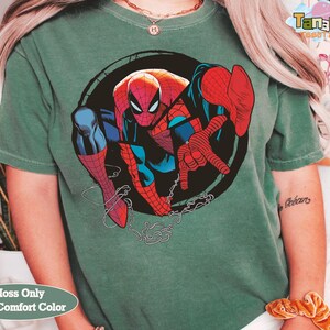 May include: Green t-shirt featuring a large graphic of Spider-Man in a dynamic pose. The superhero is depicted in his classic red and blue suit, set against a black circular background. The shirt has the text "Moss Only With Comfort Color".