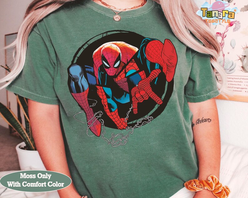 May include: Green t-shirt featuring a large graphic of Spider-Man in a dynamic pose. The superhero is depicted in his classic red and blue suit, set against a black circular background. The shirt has the text "Moss Only With Comfort Color".