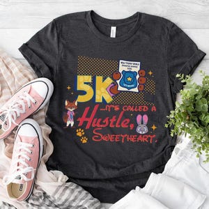 May include: Dark gray t-shirt with a colorful graphic that reads "5K... It's Called a Hustle, Sweetheart." The design includes cartoon characters, paw prints, and a police badge. Pink sneakers are next to the shirt.
