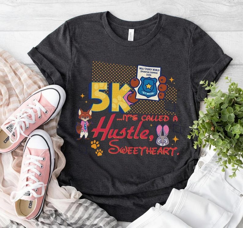 May include: Dark gray t-shirt with a colorful graphic that reads "5K... It's Called a Hustle, Sweetheart." The design includes cartoon characters, paw prints, and a police badge. Pink sneakers are next to the shirt.
