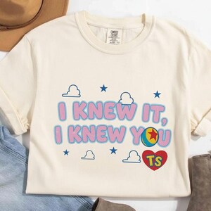 K&ouml;nnte beinhalten: Ein cremefarbenes Kurzarm-T-Shirt mit dem Text I KNEW IT, I KNEW YOU in Pink mit blauen Umrandungen. Das Design enth&auml;lt blaue Sterne, wei&szlig;e Wolken, einen bunten Ball mit einem roten Stern und ein rotes Herz mit den Buchstaben TS.