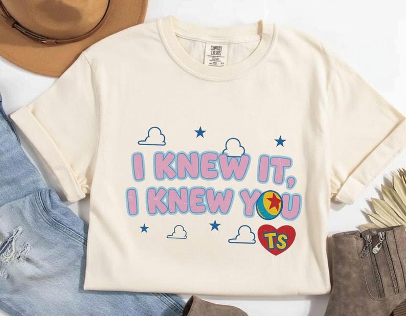 K&ouml;nnte beinhalten: Ein cremefarbenes Kurzarm-T-Shirt mit dem Text I KNEW IT, I KNEW YOU in Pink mit blauen Umrandungen. Das Design enth&auml;lt blaue Sterne, wei&szlig;e Wolken, einen bunten Ball mit einem roten Stern und ein rotes Herz mit den Buchstaben TS.