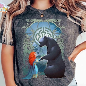 May include: Dark gray t-shirt featuring a graphic of a black bear and a girl with red curly hair. The bear and girl are in front of a blue and green background with a Celtic knot design. The shirt has short sleeves.