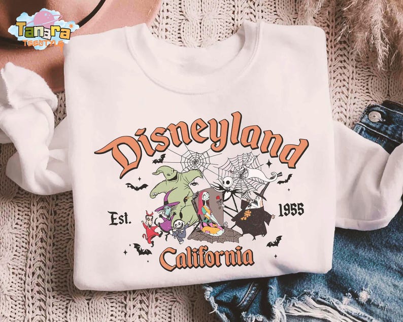 May include: A white crewneck sweatshirt featuring a graphic print with the text Disneyland Est. 1955 California. The design includes characters from The Nightmare Before Christmas, spiderwebs, and bats in orange, green, purple, and black colors.