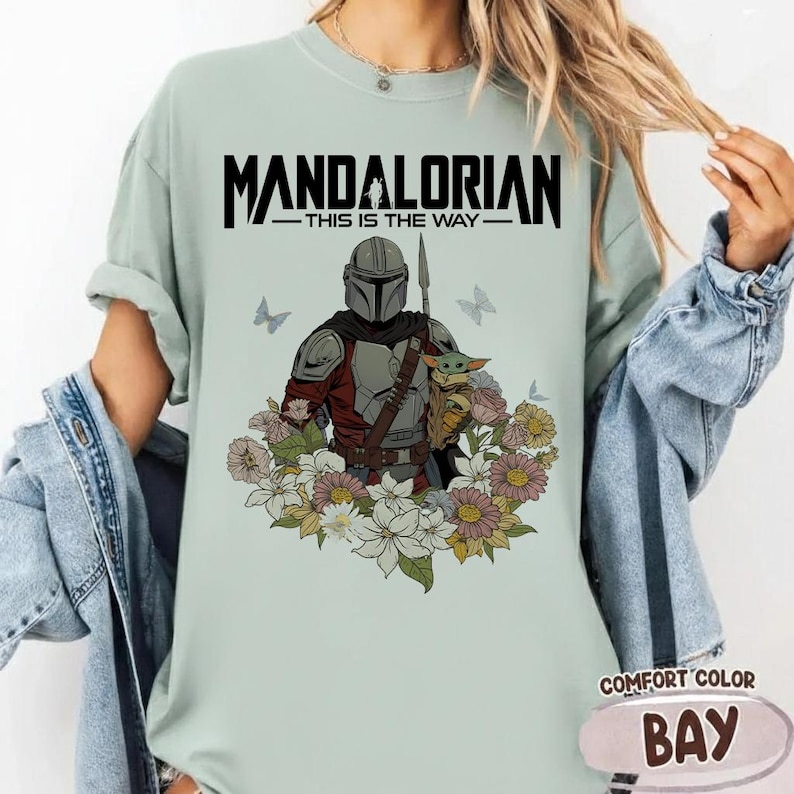 Puede incluir: Camiseta verde claro con el texto "MANDALORIAN - THIS IS THE WAY" sobre una ilustraci&oacute;n del Mandaloriano y Grogu rodeados de flores y mariposas. La camiseta es de la marca Comfort Color, color Bay.