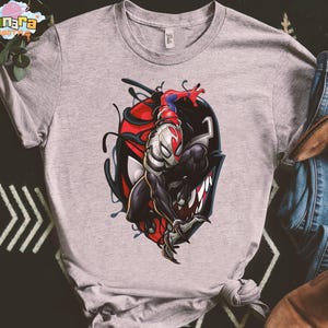 May include: A light gray t-shirt featuring a graphic of a superhero character. The design includes a red, black, and white illustration of a superhero figure. The shirt is paired with blue jeans and brown boots.