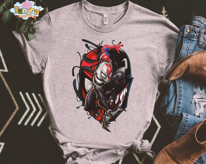 May include: A light gray t-shirt featuring a graphic of a superhero character. The design includes a red, black, and white illustration of a superhero figure. The shirt is paired with blue jeans and brown boots.