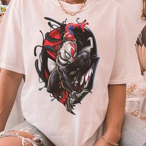 May include: White t-shirt featuring a graphic of Spider-Man merged with Venom. The design includes red, black, and white colors, with the Spider-Man figure in a dynamic pose. The shirt has short sleeves and a relaxed fit.