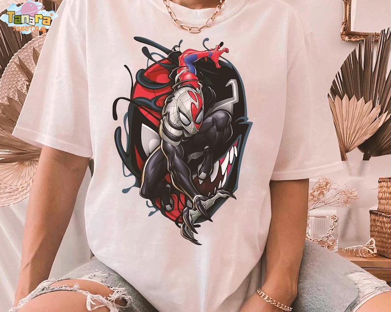 May include: White t-shirt featuring a graphic of Spider-Man merged with Venom. The design includes red, black, and white colors, with the Spider-Man figure in a dynamic pose. The shirt has short sleeves and a relaxed fit.
