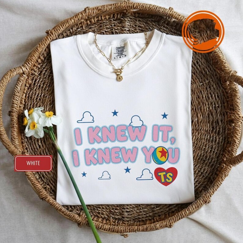 K&ouml;nnte beinhalten: Ein wei&szlig;es T-Shirt mit dem Text I KNEW IT, I KNEW YOU in Pink mit blauen Umrandungen. Das Design enth&auml;lt blaue Sterne, wei&szlig;e Wolkenumrisse, einen bunten Ball mit einem roten Stern und ein rotes Herz mit den gelben Buchstaben TS.