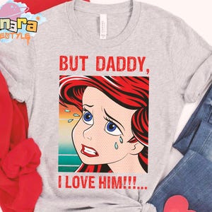 May include: Light gray t-shirt featuring a pop art style illustration of a crying cartoon character with red hair and blue eyes. The text "BUT DADDY, I LOVE HIM!!!" is printed in red.