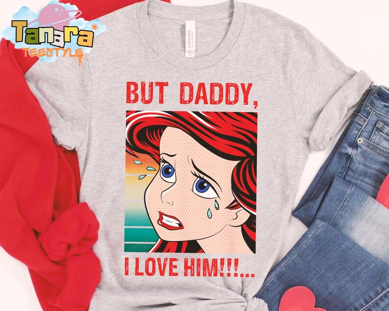 May include: Light gray t-shirt featuring a pop art style illustration of a crying cartoon character with red hair and blue eyes. The text "BUT DADDY, I LOVE HIM!!!" is printed in red.