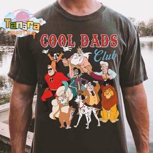 May include: Dark gray t-shirt with the text "COOL DADS Club" in red and blue. The shirt features a colorful graphic of various cartoon characters, including The Incredibles, Goofy, and Simba.