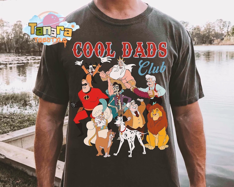 May include: Dark gray t-shirt with the text "COOL DADS Club" in red and blue. The shirt features a colorful graphic of various cartoon characters, including The Incredibles, Goofy, and Simba.