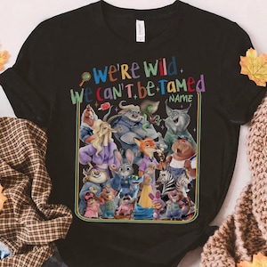 May include: Black t-shirt featuring a colorful graphic of various cartoon characters. The text "We're Wild, We Can't Be Tamed" is displayed above the characters, with a space for a name below. The design is outlined in a gold border.